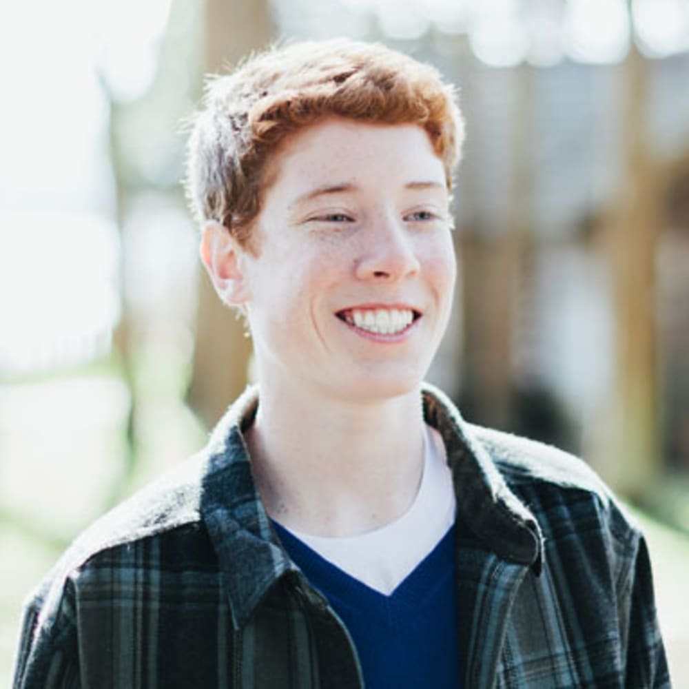 A teenage boy with ginger hair and freckles is standing outside smiling.