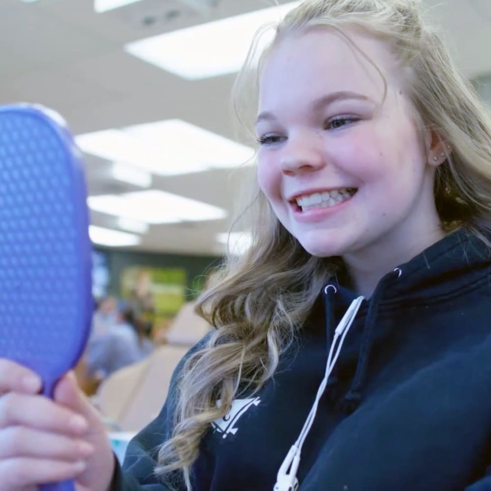 A teenage girl is sitting in the office smiling while looking at her teeth in a handheld mirror.