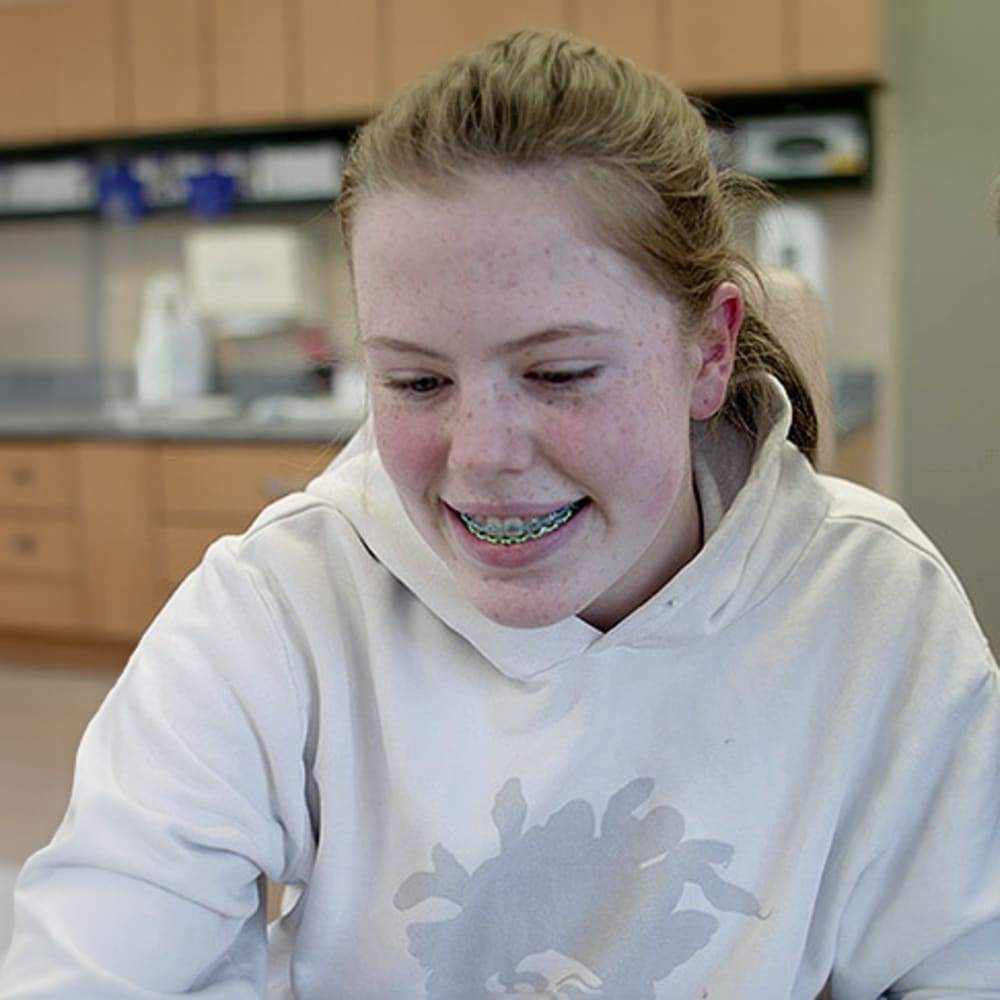 A teenage girl with red hair and freckles is sitting in the office smiling, revealing a set of braces.